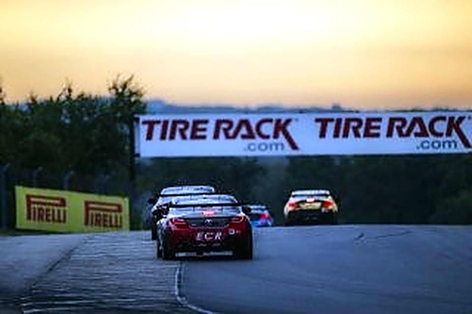 The #90 racing at sunset under a trackside banner at Road America